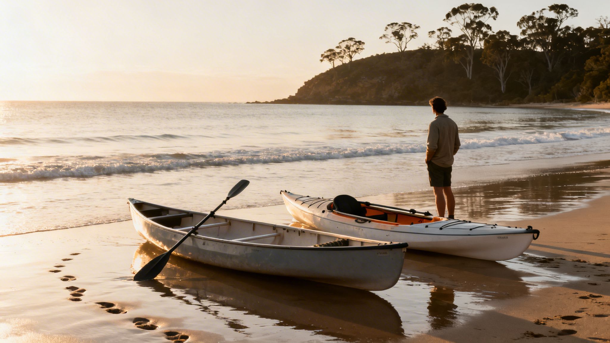 A man standing on a beach with a canoe and kayak, watching the waves at golden hour.