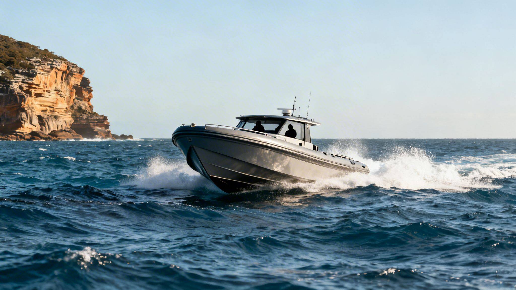 A powerful grey inflatable rescue boat speeds through choppy blue water near a rocky cliff.