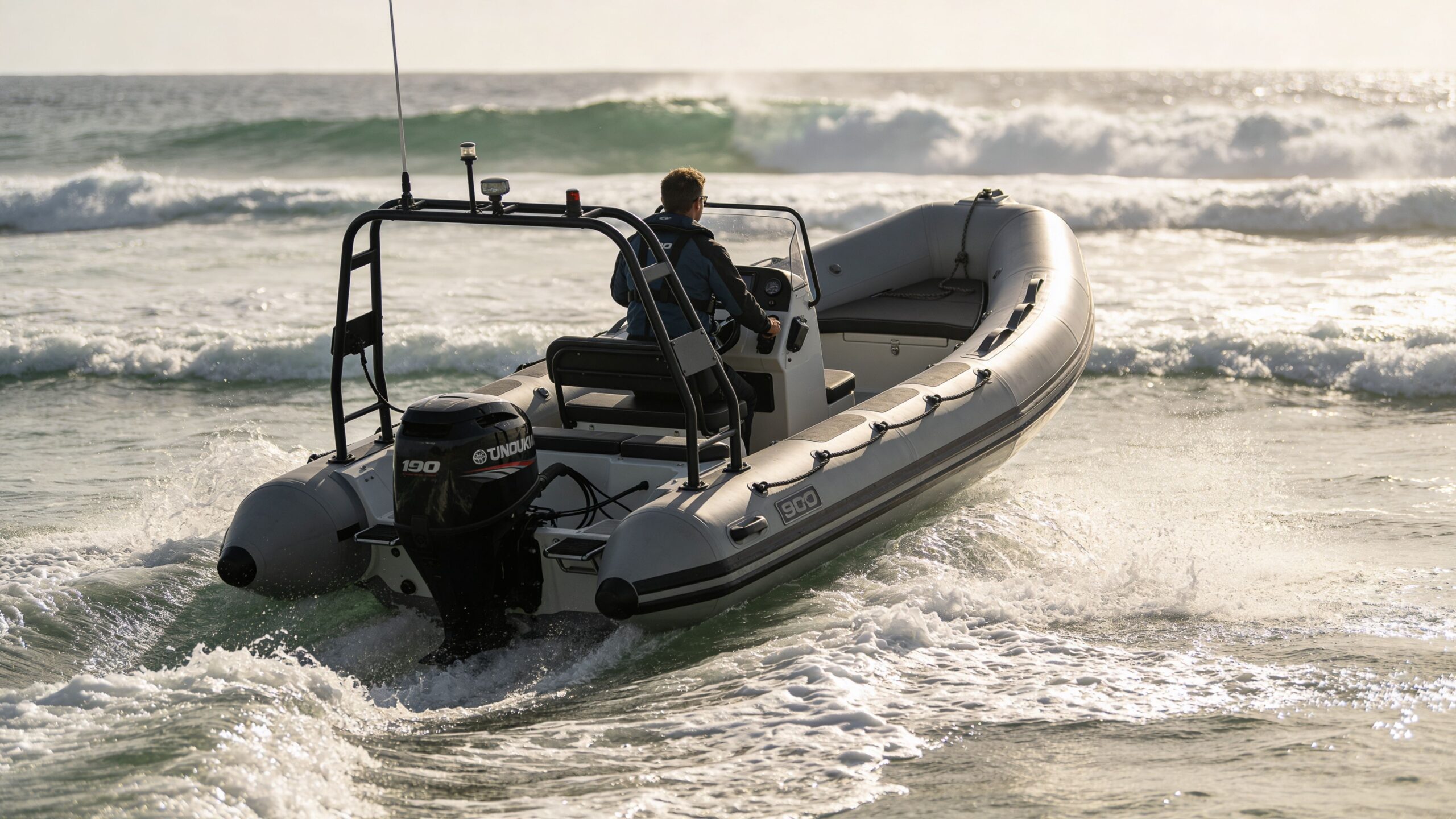 A person piloting a grey inflatable boat through ocean waves in a sunny coastal environment.