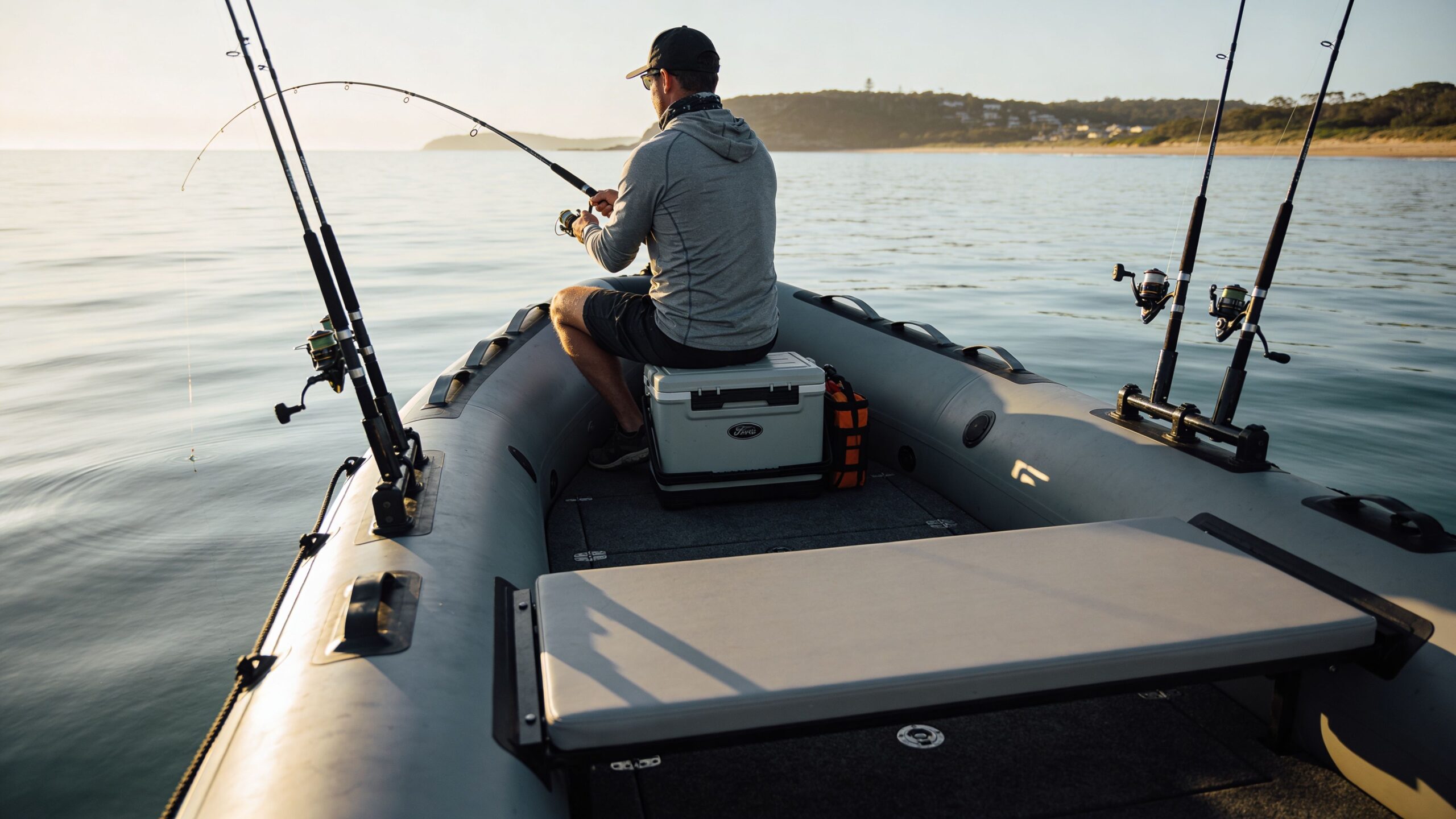 A man wearing a hat and hoodie fishes from an inflatable boat during a calm sunrise.